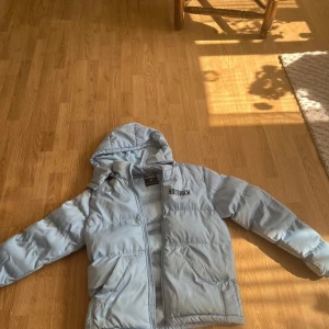 Ljusblå pufferjacka från Hoodrich - Säljer en ljusblå pufferjacka från Hoodrich med huva och dragkedja. Jackan har stora fickor framtill och Hoodrich-logga både fram och bak. Perfekt för kalla dagar och riktigt snygg streetstil.