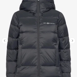 Svart pufferjacka från Sail Racing - Svart pufferjacka från Sail Racing med huva och diskret logga på bröstet. Jackan har en stilren look med breda, quiltade sektioner och dragkedja framtill. Jackan är perfekt för kalla dagar. Snygg och funktionell design som håller värmen. Jackan är använd en säsong men inga slitage eller tecken på användning.
