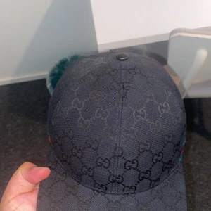 Svart Gucci keps med GG-mönster - Säljer en svart keps från Gucci med klassiskt GG-monogram över hela kepsen. Kepsen har böjd skärm och ikoniskt grönt och rött band på sidan. Tillverkad i bomullsmaterial och har justerbar rem baktill för perfekt passform. Perfekt för dig som vill ha en stilren och lyxig accessoar.