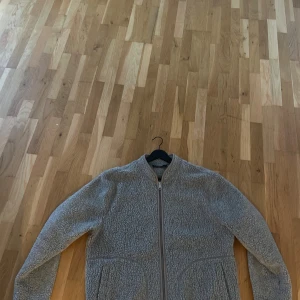 Grå teddyjacka från J.Lindeberg - Mysig grå teddyjacka från J.Lindeberg med hel dragkedja framtill och två stora fickor. Jackan har en rundad krage och är tillverkad i ett fluffigt fleece-material som håller dig varm under kyliga dagar.