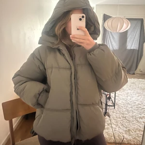 Khakifärgad pufferjacka - Säljer en khaki pufferjacka från Pull&Bear i storlek M. Jackan har huva, dragkedja framtill och två fickor med dragkedja. Även dragsko i midjan. Den är vadderad och har en boxig, oversized passform som är perfekt för kalla dagar. Tyvärr har ”ögglan” där man hänger upp jackan gått sönder. 