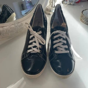 Svarta Quazi sneakers med klack - Unika svarta sneakers från Quazi i glansigt skinn med vita snören och kontrastsömmar. Skorna har rund tå, vit sula och låg klack som ger en cool twist på klassiska sneakers. Perfekta för dig som vill sticka ut med din stil.