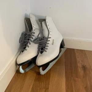 Vita skridskor Victoria med snörning - Säljer ett par klassiska vita skridskor från Victoria med grå snörning och svart klack. Skorna har högt skaft och är tillverkade i syntetmaterial med silvriga metallblad. 