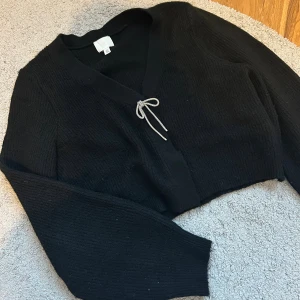 Svart croppad kofta från H&M - Svart croppad kofta från H&M med v-ringning och lång ärm. Koftan har en söt snörning framtill och är stickad i ett mjukt material. Perfekt för lager-på-lager och chill dagar.