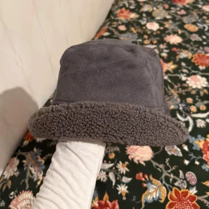 Mörkgrå bucket hat med teddyfoder - Säljer en mörkgrå bucket hat med mjukt teddyfoder runt kanten. Hatten har en klassisk form och är perfekt för kalla dagar. Materialet är syntet med en mysig, fleece-liknande känsla.