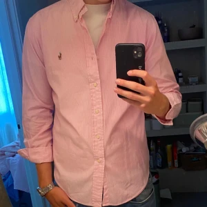 Rosa randig skjorta från Ralph Lauren - Snygg rosa skjorta från Ralph Lauren med diskreta vita ränder och klassisk button-down krage. Skjortan har lång ärm, knappar framtill och den ikoniska broderade loggan på bröstet. Tillverkad i mjuk bomull som känns skön mot huden.
