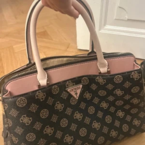 Svart och rosa handväska från Guess - Snygg handväska från Guess i svart med beige mönster och ljusrosa detaljer. Väskan har två rejäla handtag, dragkedja upptill och flera fack inuti. Tillverkad i syntetmaterial med klassiskt Guess-mönster och logga framtill.