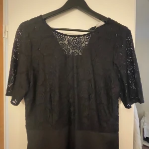 Svart spetsklänning från Dea - Svart klänning från Dea med vacker spets upptill och halvärmade ärmar. Klänningen har ett blommigt spetsmönster, rund halsringning och dragkedja i ryggen. Kjoldelen är slät och stilren, vilket ger en elegant look. Perfekt för dig som gillar detaljer och klassisk stil.