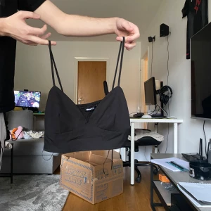 Svart croppad linnetopp från Gina Tricot - Säljer en svart croppad linnetopp från Gina Tricot med smala axelband och omlott-detalj framtill. Toppen har en v-ringad skärning och smockad rygg för extra komfort. Perfekt att styla med jeans eller kjol för en trendig look.