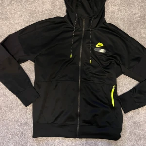Nike Air Max svart hoodie med dragkedja - Nike Air Max hoodie i svart med neongula detaljer och logga på bröstet och ryggen. Tröjan har huva med snörning, hel dragkedja och fickor framtill. Tillverkad i mjukt syntetmaterial, perfekt för träning eller chill. Snygg och sportig look.