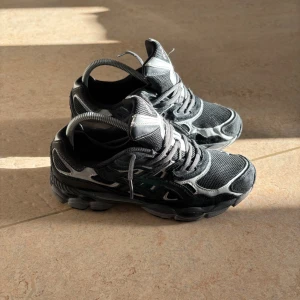 Svarta Asics sneakers - Säljer ett par svarta Asics sneakers med grå och silverfärgade detaljer. Skorna har mesh- och mockapaneler, snörning och en chunky sula som ger en sportig vibe.