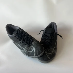 Svarta Nike fotbollsskor med snörning - Säljer ett par svarta Nike fotbollsskor med grå detaljer och snygga mönster på sidorna. Skorna har platt sula och rund tå, samt snörning för bra passform. Perfekta för inomhusspel och har en sportig vibe med stilrena linjer och Nike-logga på både sidor och sulan. Storlek 40,5.