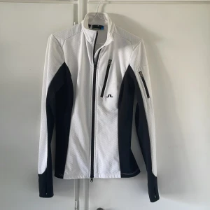 Vit och svart vindjacka från J.Lindeberg - Snygg vit och svart vindjacka från J.Lindeberg med sportig look. Jackan har lång ärm, flera dragkedjeförsedda fickor och diskret mönstrad yta. Perfekt passform och stilren design med logga på bröstet och nacken. Materialet är lätt och funktionellt.