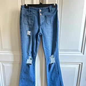 Blå bootcut jeans med slitningar mid waist - Snygga blå jeans med bootcut passform och råa, fransiga benslut. Byxorna har slitna detaljer och patchade hål på benen samt två framfickor. Stängs med dubbla knappar i midjan. Perfekta för en avslappnad och trendig look.