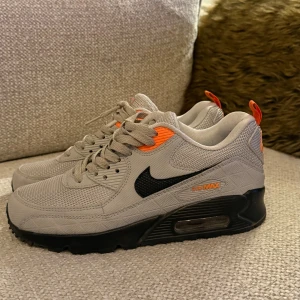 Nike Air Max 90  - Nike Air Max 90 sneakers i ljusgrå mesh och syntet med svarta detaljer och orangea accenter på häl, logga och snörningspanel. Klassisk rund tå, platt sula med synlig Air Max-dämpning och snörning. Perfekt för dig som gillar streetstyle och sportig vibe.
