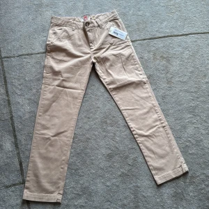 Aldrig använda beiga chinos från American Outfitters - Snygga beiga chinos från American Outfitters med klassisk rak passform. Byxorna har fickor fram, bälteshällor och stängs med knapp och dragkedja. Perfekta för en clean och stilren look. Materialet är mjukt och skönt bomullstyg.