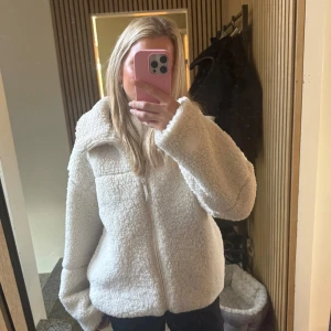 Vit teddyjacka med dragkedja - Säljer en fluffig vit teddyjacka med hög krage och dragkedja framtill. Jackan har en oversized passform och är tillverkad i mjukt fleece-material som ger en mysig vibe. Perfekt för kalla dagar och enkel att styla med jeans eller leggings.