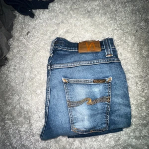 Blå jeans från Nudie Jeans - Säljer ett par klassiska blå jeans från Nudie Jeans med snygga slitningar och deras ikoniska broderade bakficka. Jeansen har normal passform och är tillverkade i mjukt denimtyg med kontrastsömmar och läderpatch i midjan. Skriv för mer bilder 
