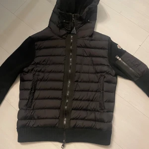 Svart Moncler cardigan - Snygg svart dunjacka från Moncler med stickade ärmar och huva. Jackan har dragkedja framtill, ribbade muddar och en cool ficka med dragkedja på ärmen. Perfekt för dig som vill ha en stilren och varm jacka med exklusiv känsla. Hör gärna av er vid intresse😀