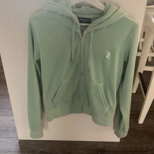 Mintgrön velourhoodie Juicy Couture M - Superfin mintgrön hoodie från Juicy Couture i mjuk velour. Tröjan har dragkedja med J-detalj, huva, känguruficka och broderad logga på bröstet. Perfekt för en chill look. 