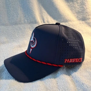Mörkblå keps med golfbrodyr - Snygg mörkblå keps från Parfect med ventilerande perforerade sidor och justerbar baksida. Framtill finns en brodyr med golfmotiv och en röd detalj-lina längs skärmen. Perfekt för golfbanan eller som cool accessoar till vardags.