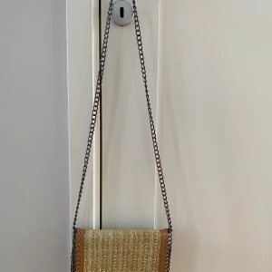 Beige axelväska med kedja och flätad front - Snygg beige axelväska med flätad front i naturmaterial och detaljer i brunt skinn. Väskan har en lång kedjerem och stängs med magnetlås. Insidan har flera fack, inklusive ett med dragkedja. Perfekt för dig som gillar en boho-inspirerad stil. Mått  Bredd: ca 23 cm  Djup: ca 3 cm  Höjd: ca 16 cm  Kommer från ett djur och rökfritt hem. 