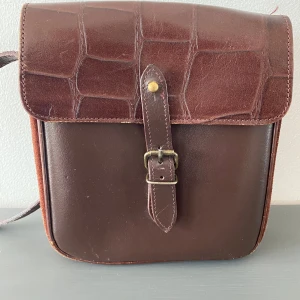 Brun axelväska i skinn med spänne - Snygg brun axelväska i skinn med krokopräglat lock och rem. Väskan har ett klassiskt spänne framtill och axelremmen har samma mönster som locket. Sidorna är i mocka vilket ger en cool kontrast till det släta skinnet. Mått  Bredd: ca 19 cm  Djup: ca 5 cm  Höjd: ca 19 cm    Kommer från ett djur och rökfritt hem. 