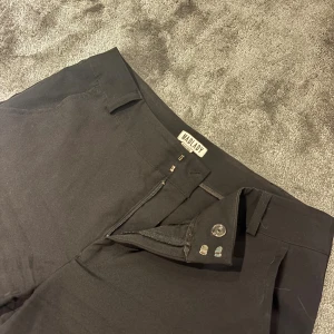 Svarta kostymbyxor från Madlady - Svarta kostymbyxor från Madlady med klassisk rak passform. Byxorna har hällor för bälte, diskreta fickor och stängs med dragkedja och hyska. Perfekta för en clean och stilren look.