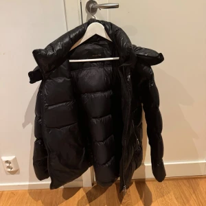 Svart pufferjacka från Massimo Dutti - Säljer en svart pufferjacka från Massimo Dutti med hög krage och dragkedja. Jackan är vadderad och har en glansig finish som ger en cool look. Perfekt för kalla dagar när du vill hålla stilen och värmen.