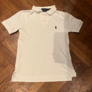 Vit pikétröja från Polo Ralph Lauren - Klassisk vit pikétröja från Polo Ralph Lauren i bomull. Barnstorlek S(8) pojke. Tröjan har korta ärmar, krage med två knappar och den ikoniska broderade loggan på bröstet. Perfekt för en clean och stilren look.