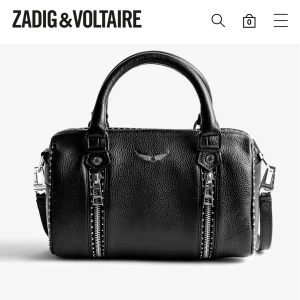 Svart handväska från Zadig & Voltaire - Säljer en cool svart handväska i skinn från Zadig & Voltaire med två rejäla handtag, silverfärgade detaljer och dragkedjor framtill.  Väskan är använd och har några synliga tecken på att den är använd. Väskan har en boxig form och ett snyggt emblem i metall på framsidan. Perfekt för dig som gillar edgy och stilren design. Köpte för ungefär 4800 kr.