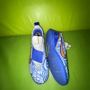 Nike CR7 inomhusskor - Säljer ett par Nike fotbollsskor med grymt blått och vitt grafiskt mönster och orange swoosh. Skorna har elastiskt band över vristen och blå sula med bra grepp. Perfekta för dig som vill sticka ut på planen!