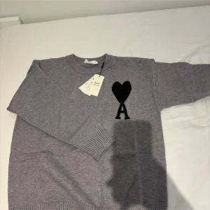 Grå stickad tröja - Snygg grå stickad tröja med svart hjärta och A-logga på bröstet. Tröjan har rund hals, ribbade muddar och är långärmad. Perfekt för dig som gillar stilrena och trendiga plagg med en twist.
