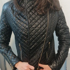 Svart quiltad skinnjacka med dragkedjor - Snygg svart quiltad skinnjacka med guldiga dragkedjor framtill, vid fickor och ärmslut. Jackan har figurnära passform och coolt rutmönster över hela ytan. Perfekt för dig som vill ha en edgy och trendig look. Har inte kollat varumärke men kan kolla ifall. Pris kan diskuteras😊
