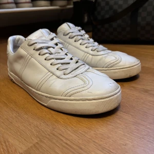 Valentino vita sneakers med nitar - Snygga vita sneakers från Valentino med coola silverfärgade nitar på hälen. Skorna är tillverkade i mjukt skinn och har klassisk snörning samt rund tå. Perfekt för dig som gillar stilrena och exklusiva sneakers med en edgy detalj. Observera att en av nitarna på hälen har lossnat, men detta är enkelt att åtgärda av en skomakare eller med lite pyssel hemma. Även snörningen vid tån kan enkelt fixas hos en skomakare vid behov. Skorna är i övrigt i mycket fint skick och har många användningar kvar! 