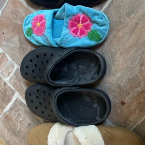 Fyra par olika stilar och storleker tofflor  - Säljer fyra par tofflor och sandaler: ett par blå plyschtofflor med broderade rosa blommor, ett par svarta syntettofflor med hål, ett par svarta tofflor med brun kant och ett par bruna tofflor med vit fluffig insida. Perfekta för att chilla hemma eller snabbt hoppa i.