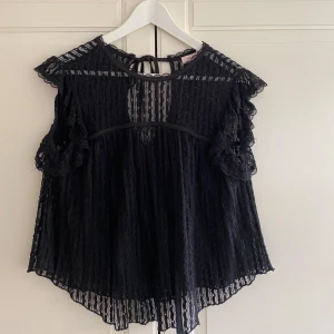 Svart spetsblus från Nelly - Supersöt svart blus från Nelly i transparent spets med volangdetaljer vid ärmarna och knytning i nacken. Blusen har en lös passform och är ärmlös med fina spetskanter. 