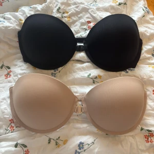 Svart och beige axelbandslösa push-up bhar - Två axelbandslösa bhar i svart och beige med avtagbara axelband. Båda har vadderade kupor och smidig knäppning fram och är justerbar baktill. Perfekt för outfits där du vill ha en clean look utan synliga axelband.
