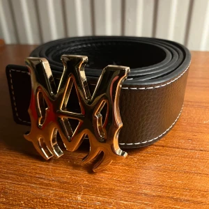 Amiri bälte - Stilrent svart Amiri bälte i skinn med markerade vita sömmar och ett stort, guldigt spänne. Bältet har en grov struktur och passar perfekt för att lyfta din outfit med en cool detalj. Helt nytt!!