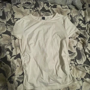 Vit t-shirt med rynkade sidor från SHEIN - Vit t-shirt från SHEIN i storlek M. Modellen har korta ärmar och rund halsringning. Sidorna är snyggt rynkade för en cool detalj. Perfekt för dig som gillar enkel och clean stil med en twist.