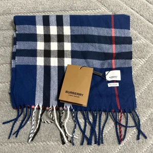 Blårutig kashmirhalsduk från Burberry - Exklusiv halsduk från Burberry i 100% kashmir, tillverkad i Skottland. Klassisk rutig design i blått, vitt, svart och röda detaljer. Mjuka fransar i ändarna och ikonisk Burberry-tag. Perfekt accessoar för att lyfta din stil.