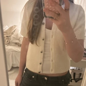 Kräm Vit kofta från Gina Tricot - Kort, vit/beige kofta från Gina Tricot med ribbade kanter och snygga knappar framtill. Koftan har korta ärmar och är gjord i ett mjukt, stickat material. Tröjan är inte stickig alls 🩷 köpte för ca 300kr 🙌