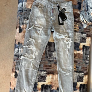 Dsquared2 ljusgrå distressed jeans - Snygga ljusgrå jeans från Dsquared2 med slitna detaljer, trasiga partier och färgstänk för en rå streetlook. Klassisk femficksmodell med dragkedja och Dsquared2-logga vid fickan. Tillverkade i bomull med en slim passform.