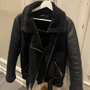 Svart fodrad skinnjacka med bälte Zara - Snygg skinnjacka från Zara med detaljer i skinn och bred krage. Jackan har dragkedja framtill, två snedställda fickor med dragkedja och ett bälte nedtill. Perfekt för kalla dagar när man också vill vara snygg. 