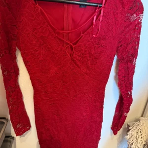 Röd spetsklänning med snörning - Röd figurnära klänning i spets med lång ärm och v-ringning framtill. Snygg snörningsdetalj vid urringningen och dragkedja bak. Perfekt för dig som vill sticka ut med en elegant och feminin look.