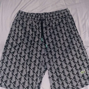 Svarta mönstrade shorts från Lacoste - Svarta shorts från Lacoste med vitt geometriskt mönster och klassisk krokodillogga broderad på benet. De har elastisk midja med snörning och är gjorda i mjuk bomull med lite stretch för extra komfort. Perfekta för chill dagar och snygga till sneakers.