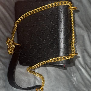 Gucci axelväska med guldkedja och pärldekor - Gucci axelväska i svart skinn med präglat GG-mönster, bred guldkedja och läderrem. Framsidan har gröna och röda paneler samt en ikonisk pärlprydd bi-detalj i guld. Insidan är vinröd och väskan stängs med ett snyggt spänne.