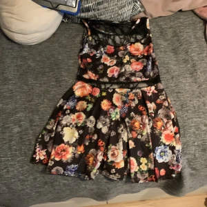 Blommig kortklänning med spetsdetalj - Supersöt kortklänning med svart botten och färgglatt blommönster i rosa, rött, gult och blått. Klänningen har ärmlös design och en snygg spets upptill som ger en extra touch. Kjolen är lätt utställd och materialet känns mjukt och följsamt mot kroppen.