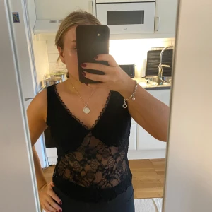 Svart spetsig linnetopp med v-ringning - Säljer en svart ärmlös linnetopp i spets med v-ringning och fina spetsdetaljer längs kanterna. Toppen är genomskinlig med blommigt spetsmönster och har en croppad passform. Perfekt för dig som vill ha en snygg och trendig look. Helt ny