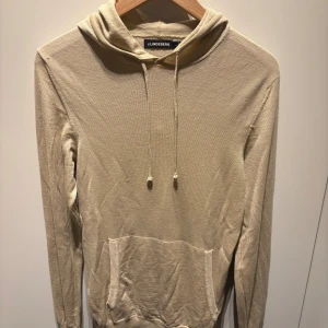 Beige hoodie med huva och ficka J.Lindeberg - Snygg beige hoodie med huva och dragsko från J Lindeberg. Tröjan har en klassisk känguruficka framtill och ribbade muddar vid ärmslut och nederkant. Perfekt för chill dagar och enkel att matcha med jeans eller joggers.